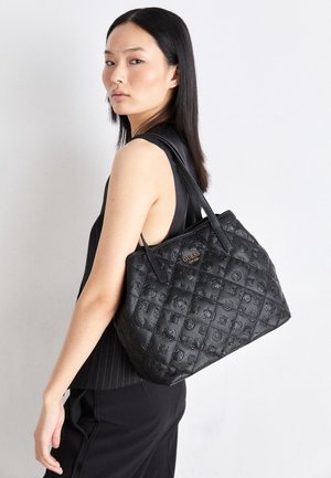 Frau mit glatten schwarzen Haaren, die ein schwarzes ärmelloses Oberteil trägt und eine große gesteppte schwarze Guess-Ledertasche an der Schulter trägt.