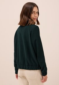 Donkergroene bomberjack met ribgebreide manchetten en zoom, gemaakt van zacht materiaal, met een relaxte pasvorm en een minimalistisch ontwerp.