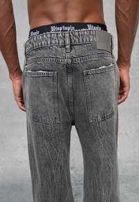 Vaqueros de mezclilla gris desgastados con un corte relajado, dos bolsillos traseros y una cinta de cintura negra con la marca "Utopía" en texto blanco.