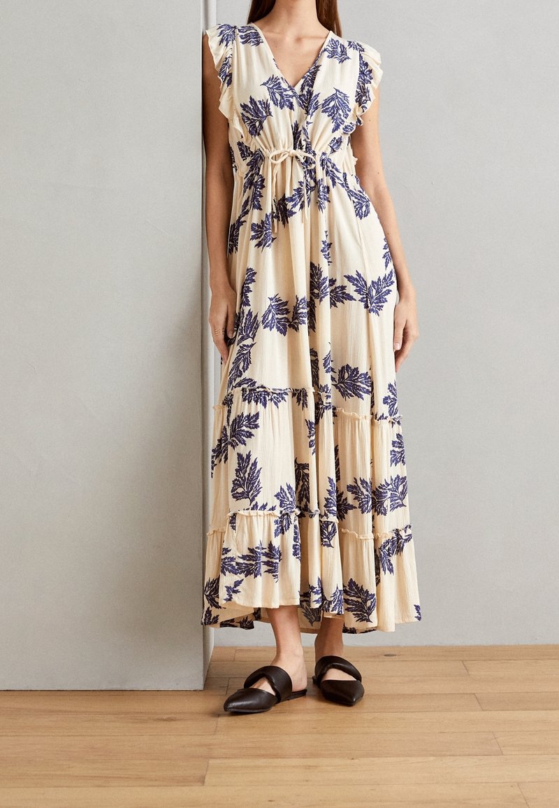 Femme portant une robe maxi crème sans manches avec des imprimés de feuilles violettes et un ourlet à volants, associée à des chaussures plates pointues noires.