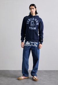 Navy hoodie with "ICECREAM TEAM" graphic, blue denim jeans featuring a white animal design, and brown shoes with pink accents.