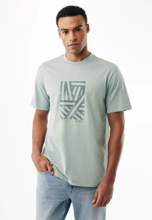 Man draagt een lichtgroen T-shirt met opdruk en lichtblauwe jeans, staat met één hand in zijn zak tegen een witte achtergrond.