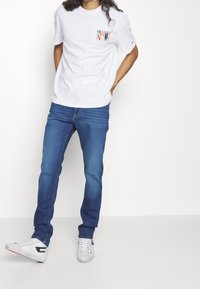 Vit kortärmad t-shirt med flerfärgad "TOMMY"-logotyp på bröstet, matchad med blå denimjeans och vita sneakers med svarta accenter.