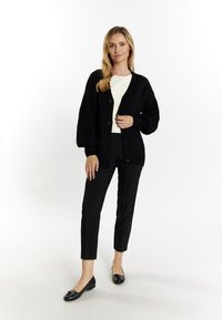 Cárdigan negro de punto con mangas texturizadas, cierre de botones, usado sobre una blusa blanca, combinado con pantalones ajustados negros y zapatos planos.