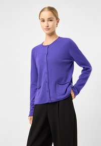 Kvinde står med hånden på hoften iført en lilla knapåbnet cardigan med lommer og sorte brede bukser foran en ensfarvet baggrund.