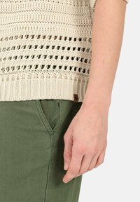 Beige gebreide trui met opengewerkte structuur, geribbelde onderkant; gecombineerd met groene broek met zijzak en rechte pijpen.