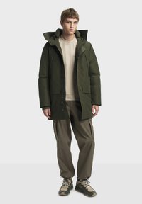 Doudoune vert olive avec capuche, dotée d'une fermeture éclair sur le devant, de deux poches poitrine et de manches longues, assortie à un pantalon cargo beige et des bottes de randonnée.