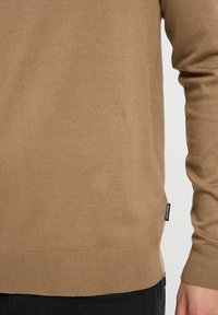 Suéter marrón de punto con mangas largas, puños y dobladillo acanalados, que presenta una pequeña etiqueta en el costado. Textura suave, corte regular.