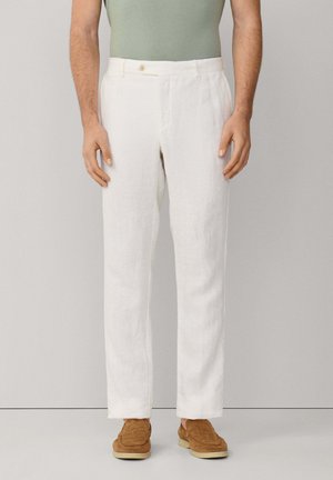 Homme portant un pantalon en lin blanc, une chemise vert clair rentrée, et des mocassins en daim marron, debout devant un fond neutre.