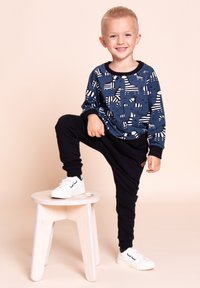 Uśmiechnięty młody chłopiec ubrany w niebieski sweter w zebrę i czarne spodnie, stojący z jedną stopą na jasnym, drewnianym stołku na beżowym tle.