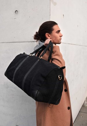 Black canvas duffel bag with leather accents, featuring dual handles and two front stripes. Worn by a person in a brown coat against a concrete wall.