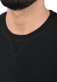 Czarna teksturowana bluza z prążkowanym, okrągłym dekoltem i subtelnym przeszyciem w kształcie litery V przy kołnierzu. Widok z bliska.