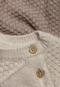 Beige stickad tröja med texturerat mönster, med en ribbad krage och två träknappar på sidan. Bakgrunden är ett dämpat taupe-tyg.