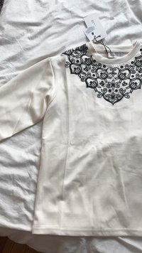 Top branco de mangas longas com intrincados bordados pretos ao longo do decote. O tecido é suave com um leve brilho. Etiquetas anexadas.