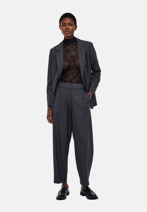 Femme debout portant une veste de costume gris foncé et un pantalon large taille haute sur un haut en dentelle noire transparente et des mocassins noirs.