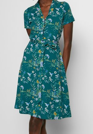 Robe bleu sarcelle longueur genou avec manches courtes, devant boutonné, ceinture nouée à la taille, et motif floral et d'oiseaux portée par une personne debout.