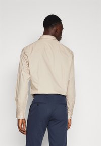 Beige Hemd mit langen Ärmeln, mit einem schlichten Rückendesign. Getragen mit navyblauen Hosen, die zwei Gesäßtaschen haben.