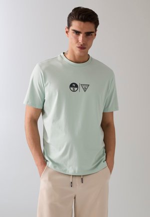 Giovane uomo con capelli scuri che indossa una t-shirt verde chiaro con loghi neri e pantaloni beige, in piedi con le mani in tasca davanti a uno sfondo semplice.