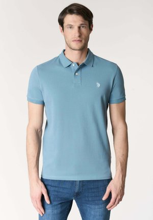Uomo che indossa una polo a maniche corte azzurro chiaro con un piccolo logo bianco sul petto sinistro e jeans blu, in piedi davanti a uno sfondo semplice.