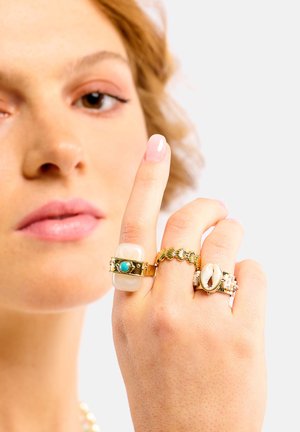 Bagues en or et en argent avec des designs variés : une grosse pierre blanche avec turquoise, une chaîne délicate, et un incrusté de coquillage sur une bague.