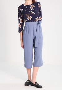 Blusa a maniche lunghe navy con fiori beige, abbinata a pantaloni cropped azzurro chiaro a vita alta e mocassini neri con dettagli dorati.