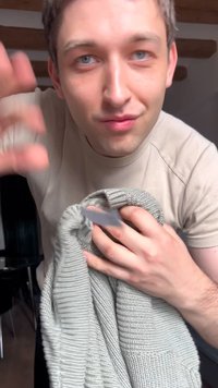 Jeune homme aux yeux bleus tenant un pull en maille gris, inspectant ou montrant son étiquette à l'intérieur, sous des poutres en bois.