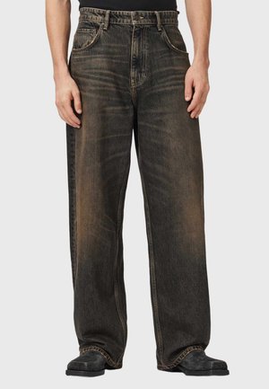 Mand iført sorte brede falmede jeans og sorte sko, der står med hænderne afslappet langs siden mod en ensfarvet baggrund.