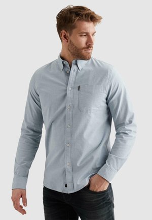 Moški z brado v svetlo modri srajci s srajčnimi gumbi in črnih hlačah, stoji z roko v žepu, gleda v stran pred enobarvnim ozadjem.