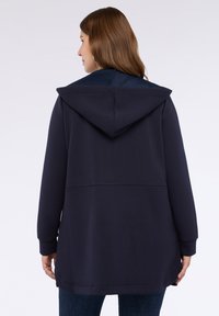Žena s dlouhými hnědými vlasy má na sobě námořnický modrý oversize svetr s kapucí staženou dolů, v kombinaci s tmavě modrými džíny, zobrazená zezadu na bílém pozadí.