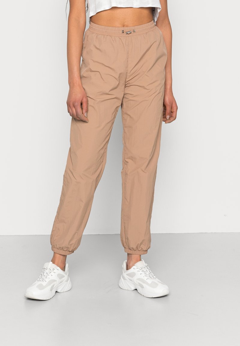 Personne portant un pantalon de jogging beige à taille élastique et des baskets blanches épaisses, debout devant un fond gris clair uni.