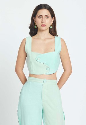 Femme aux longs cheveux foncés porte un crop-top bleu pastel avec de gros boutons et un pantalon taille haute assorti, debout sur un fond blanc.