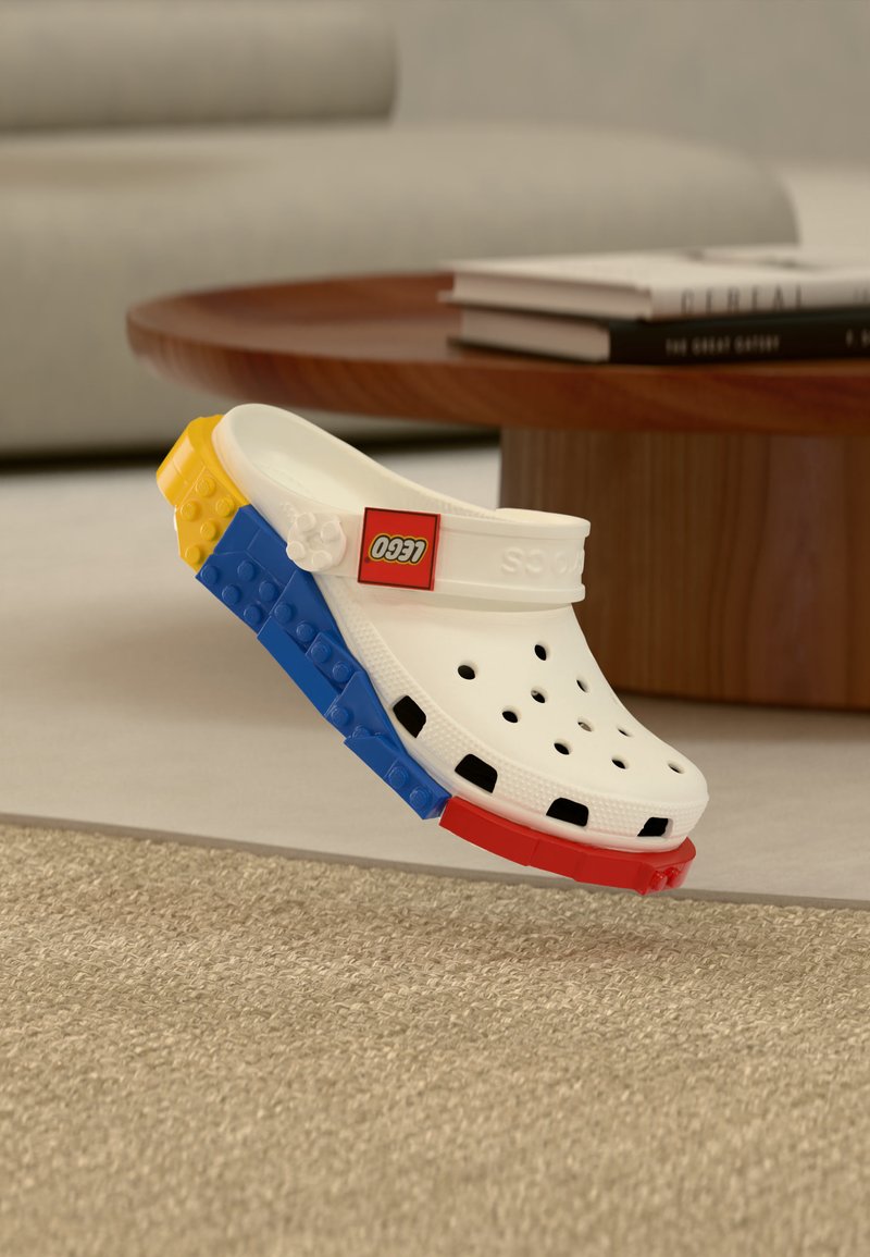 Weißer Clog mit von LEGO-Steinen inspirierter Sohle in Rot, Blau und Gelb, mit Belüftungslöchern und rotem LEGO-Logo auf dem Riemen.