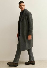 Cappotto in lana grigio con colletto a sciallato, indossato sopra una camicia scura. Abbinato a jeans grigi e stivali brown slip-on. Texture complessiva liscia.