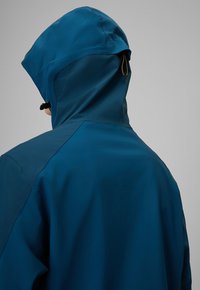 Personne portant une veste imperméable bleu foncé avec une capuche ajustable, vue de dos sur un fond gris uni.