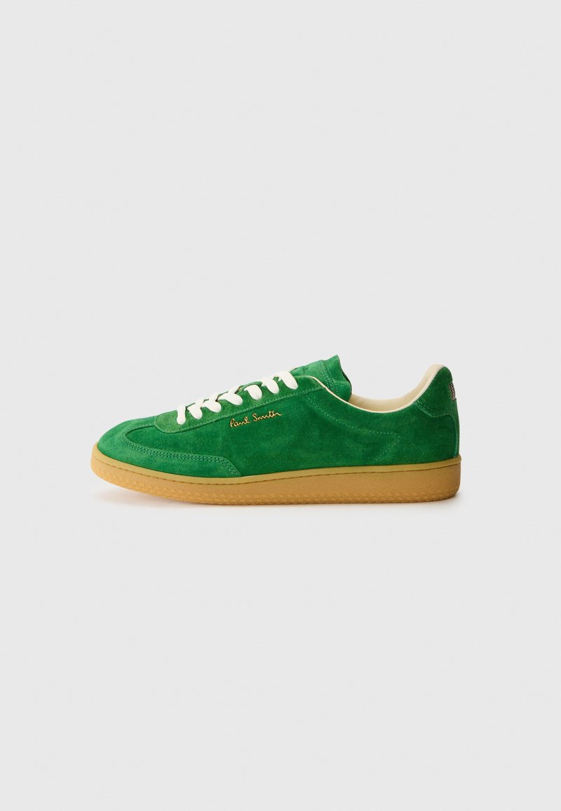 Baskets en suède vert avec une semelle en caoutchouc, des lacets blancs et un logo doré sur le côté. Texture lisse avec des détails de couture distincts.
