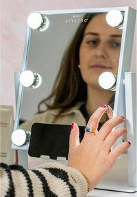Spegelskåp med justerbara LED-lampor, elegant vit ram och smartphonehållare. Har en reflekterande yta och användare som justerar ljuset.