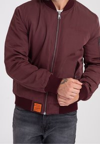 Burgundy bomberjacka med ribbad mudd, framsida med dragkedja, sidofickor och en fyrkantig orange etikett vid nederkanten. Slät, något blinkande tyg.