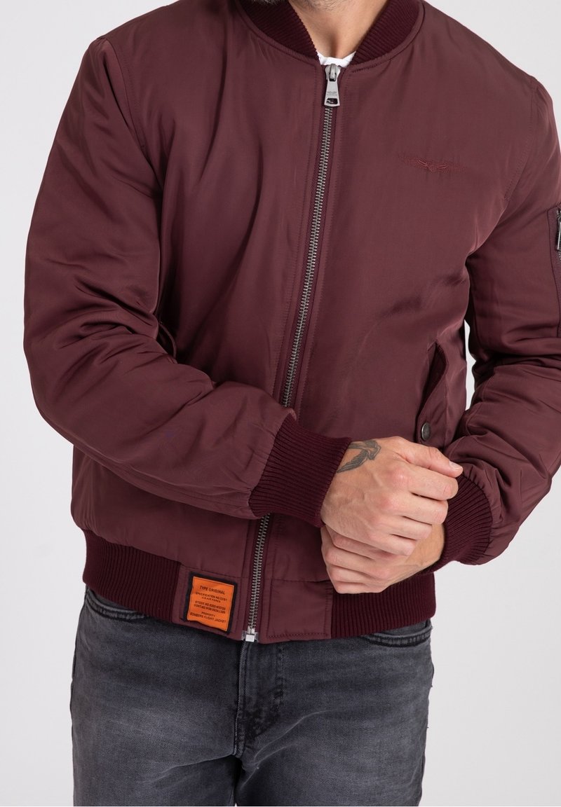 Burgundy bomberjacka med ribbad mudd, framsida med dragkedja, sidofickor och en fyrkantig orange etikett vid nederkanten. Slät, något blinkande tyg.