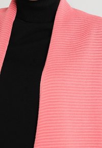 Różowy kardigan prążkowany z otwartym przodem, noszony na czarnym swetrze z golfem. Kardigan posiada miękką fakturę i delikatne poziome paski.