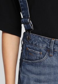 Jeansoverall med justerbar rem fästad med metallspänne, bärs över en svart t-shirt med kort ärm, närbild på midjan och vänster arm.