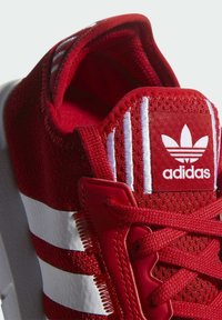 Czerwony but sportowy z siateczkową cholewką, z białymi paskami, gumową podeszwą i haftowanym logo na języku. Teksturowany obszar sznurowania.