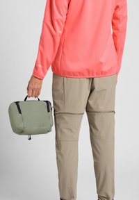 Borsa leggera verde con zip e manico nero, tenuta da una persona che indossa una camicia a maniche lunghe corallo brillante e pantaloni beige.