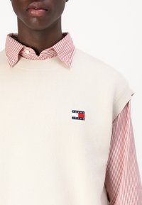 Krémový pletený bezrukávový svetr přes červenou a bílou pruhovanou košili, se malým námořnickým a červeným logem "TOMMY JEANS" na hrudi.