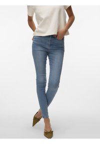 Femme portant un jean skinny bleu clair, un haut blanc à manches courtes, et des ballerines vert olive pointues avec des nœuds, debout les mains dans les poches.