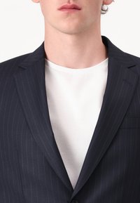 Nuori mies pukeutunut tummansiniseen liituraitapukuun ja yksinkertaiseen valkoiseen pyöreäkauluspaitaan, kuvattu nenän alapuolelta.