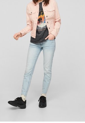 Donna con giacca rosa chiaro, t-shirt nera con stampa, jeans skinny azzurri, sneakers nere e calzini bianchi su uno sfondo grigio semplice.