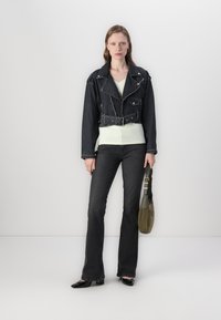 Svart kort denimjakke med glidelås, hvit topp, mørke utsvingte jeans og en grønn veske. Modellen står foran en ensfarget bakgrunn.