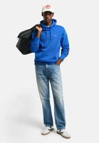 Blauwe hoodie met koordjes, zwarte tas, lichtblauwe spijkerbroek en witte sneakers met zwarte accenten. Pet met 'GANT' in rode lettering.