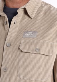 Camisa de pana beige con cuello abotonado y parche del logo Oxbow. Presenta dos bolsillos frontales con cierres de botón y textura de acanalado vertical.