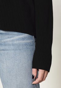 Schwarzer gerippter Pullover mit langen, weiten Ärmeln, kombiniert mit hellblauen Jeans. Der Stoff hat eine strukturierte Oberfläche.